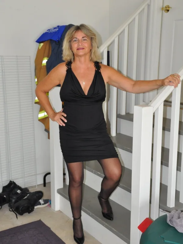 Femme en robe noire posant sur un escalier intérieur, regardant l'objectif, dans un cadre domestique.