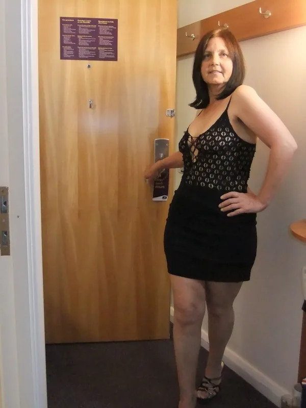 Femme en robe noire posant dans un couloir d'hôtel près d'une porte, avec une salle de bain visible à gauche.