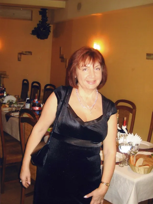 Femme souriante en robe noire dans un restaurant, regardant l'objectif, avec des tables dressées en arrière-plan.