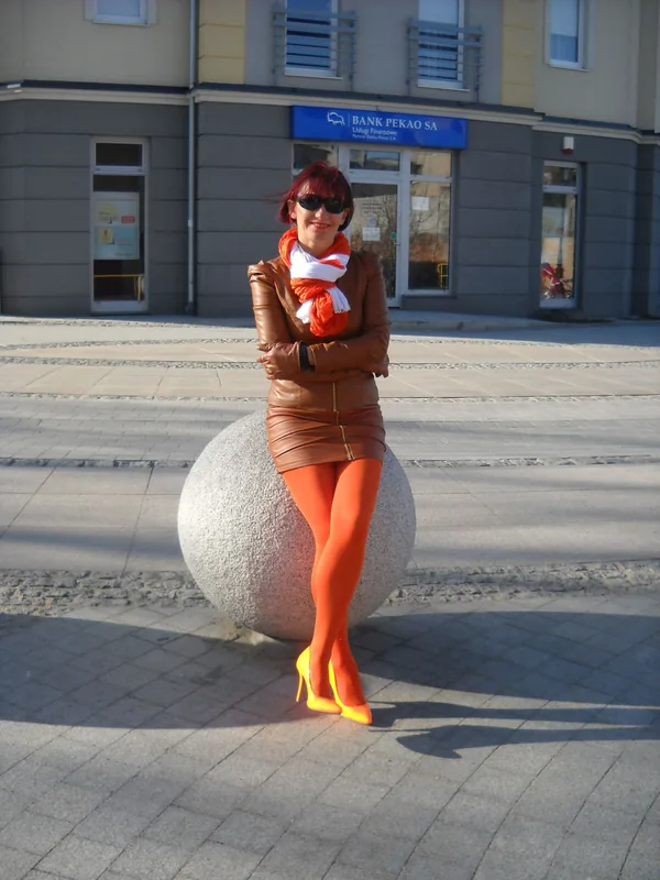 Femme aux cheveux roux en tenue cuir marron, collants orange, lunettes de soleil, assise sur une sphère en pierre dans une rue urbaine.
